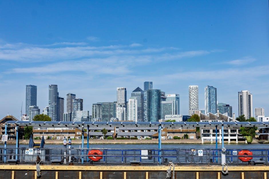 A view of a modern city skyline across the river, with tall skyscrapers featuring glass facades reflecting sunlight, set against a bright blue sky with scattered clouds. In the foreground, there is a