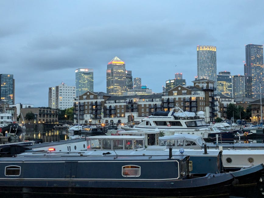 A view of a modern city skyline across the river, with tall skyscrapers featuring glass facades reflecting sunlight, set against a bright blue sky with scattered clouds. In the foreground, there is a