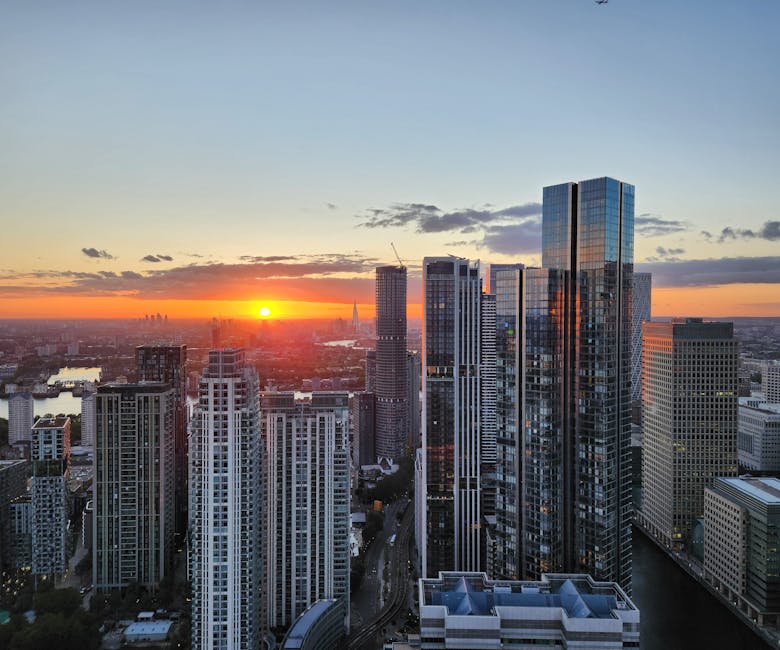 A panoramic view of a modern urban cityscape at sunset, featuring a cluster of tall skyscrapers with reflective glass facades, some under construction with cranes visible. The foreground includes vari